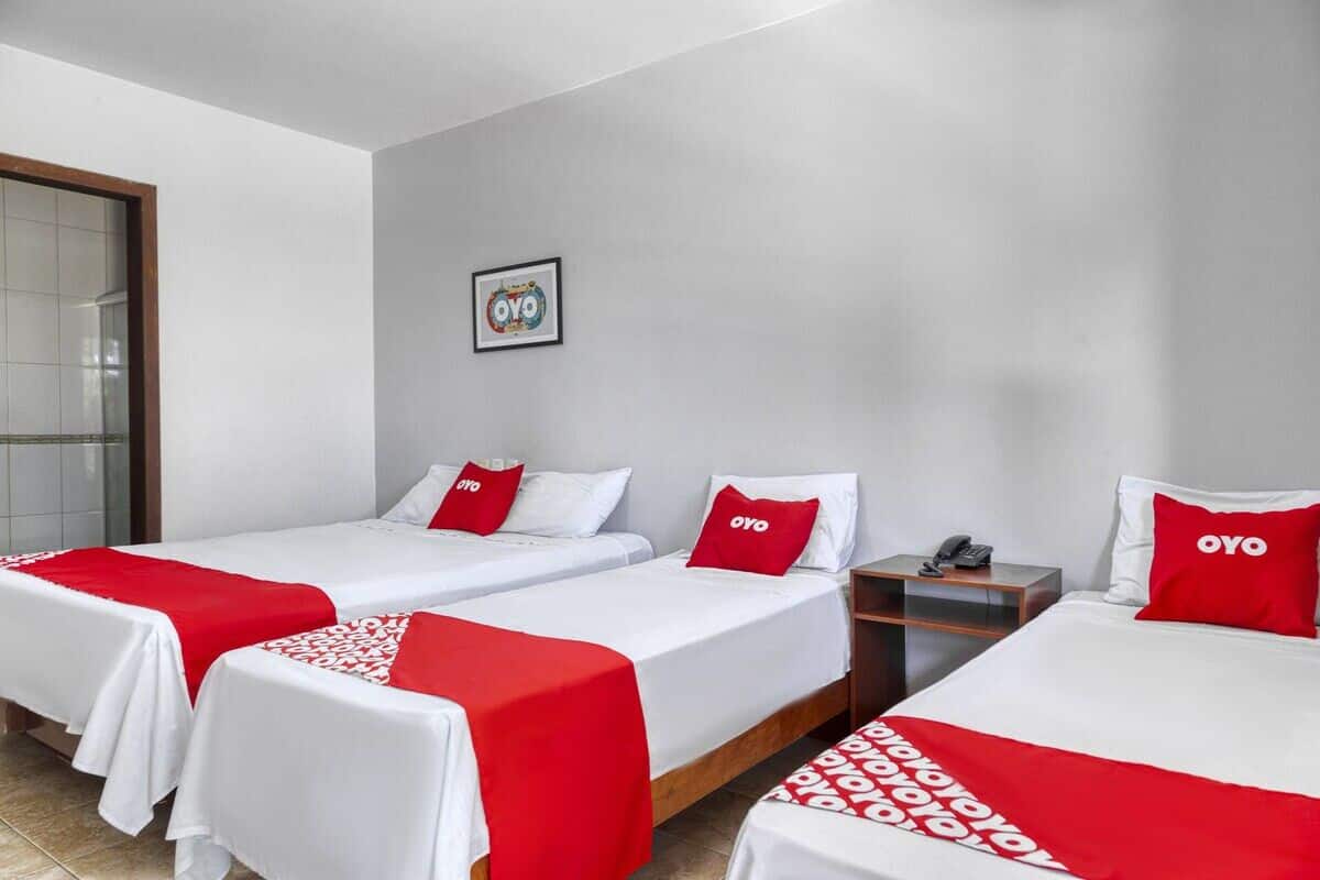 A hotel room with three beds, each with a red and white bedspread and pillows, a small bedside table with a phone, and a framed picture on the wall.