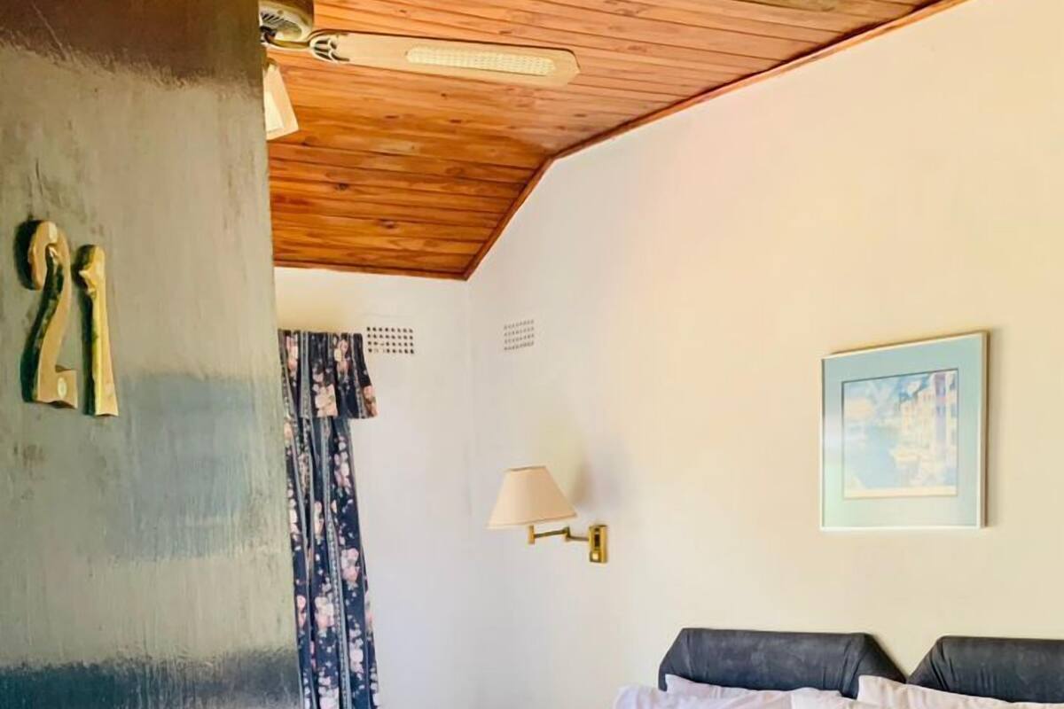 A hotel room with a wooden ceiling, a bed with white linens, a nightstand, a wall-mounted lamp, and a painting.