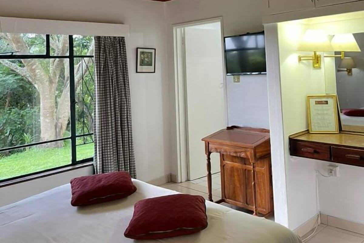 A bedroom with a wooden ceiling, a bed with two red pillows, a desk, a TV, and a window with curtains.