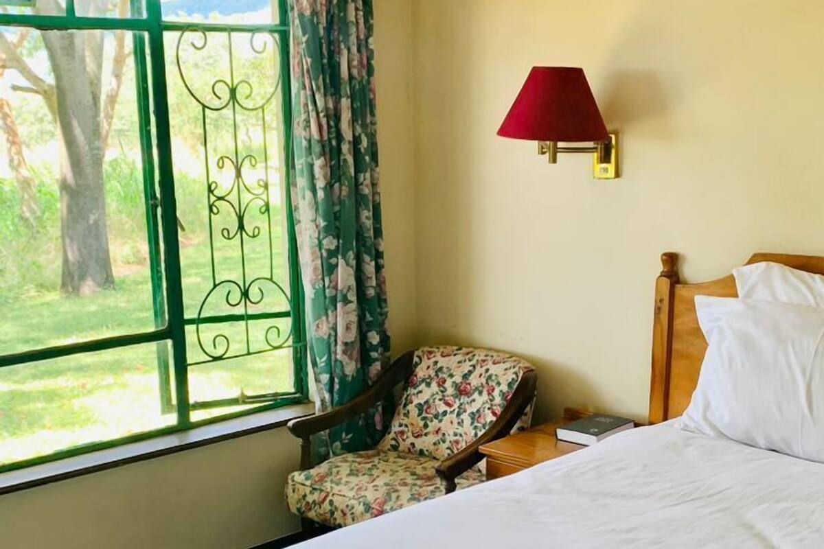 A bedroom with a large bed, a chair, a window with floral curtains, and a red wall lamp.
