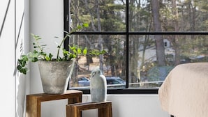 Ein Schlafzimmer mit großem Fenster, einem Bett, einer Topfpflanze und einer Vase auf einem Holztisch.