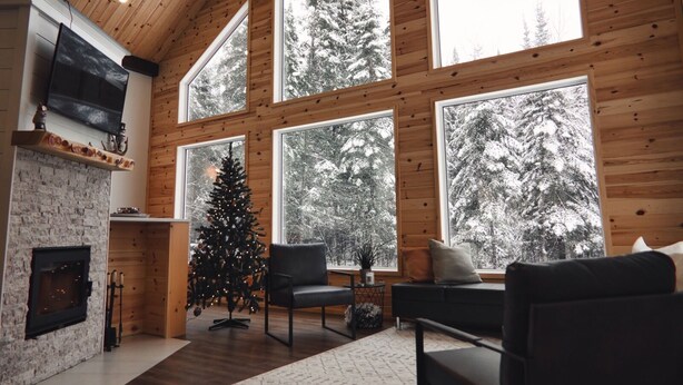 L’intérieur chaleureux d’un chalet avec une cheminée, un sapin de Noël et de grandes fenêtres donnant sur une forêt enneigée.