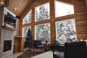 L’intérieur chaleureux d’un chalet avec une cheminée, un sapin de Noël et de grandes fenêtres donnant sur une forêt enneigée.