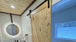 Une salle de bain moderne avec une porte coulissante en bois, un miroir rond, un meuble-lavabo avec un pare-éclaboussures en marbre et un banc près de la fenêtre.