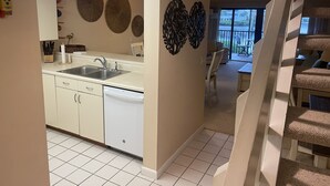 A modern kitchen with white cabinets, a dishwasher, and a large window. There are decorative wall art pieces and a dining area with a table and chairs. - 2/2.5 Waterfront condo with pools and tennis courts (Bokeelia)