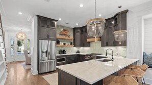 A modern kitchen with dark cabinetry, a large island, stainless steel appliances, and decorative lighting. - "Seaglass" Watersound home with Golf Cart, Pool, and Private Beach (Santa Rosa Beach)