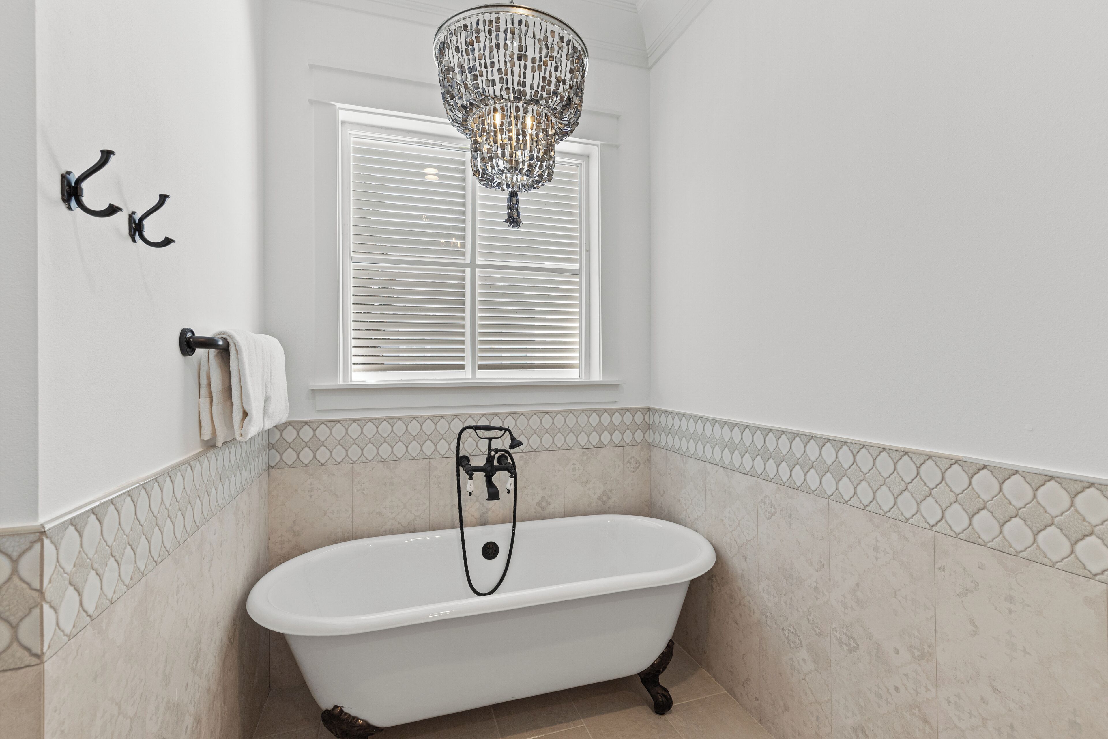 Une salle de bain avec une baignoire sur pieds, un lustre, une fenêtre avec des stores et un porte-serviettes.