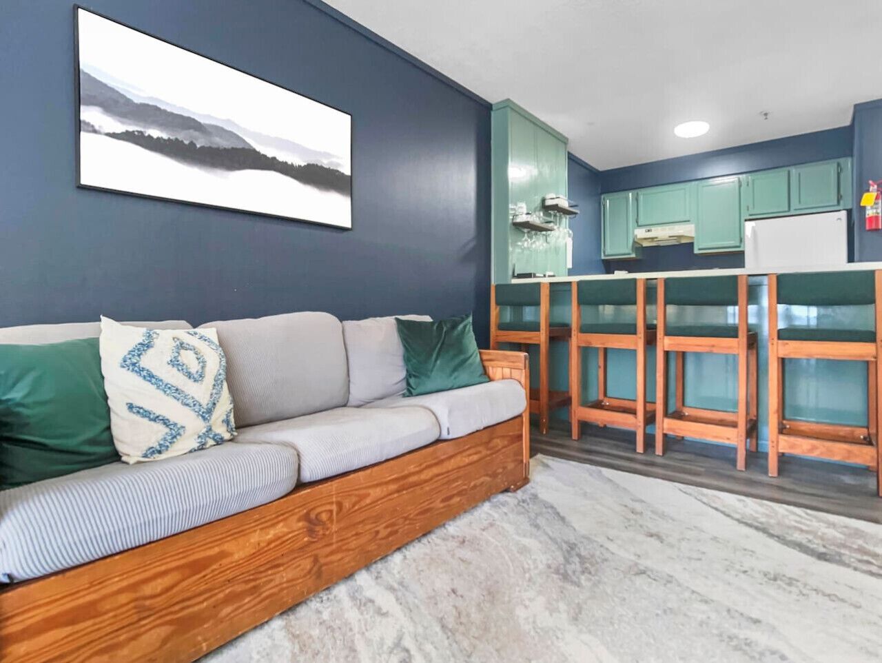 A modern living room with a wooden sofa, a kitchen area with teal cabinets, and a large window.