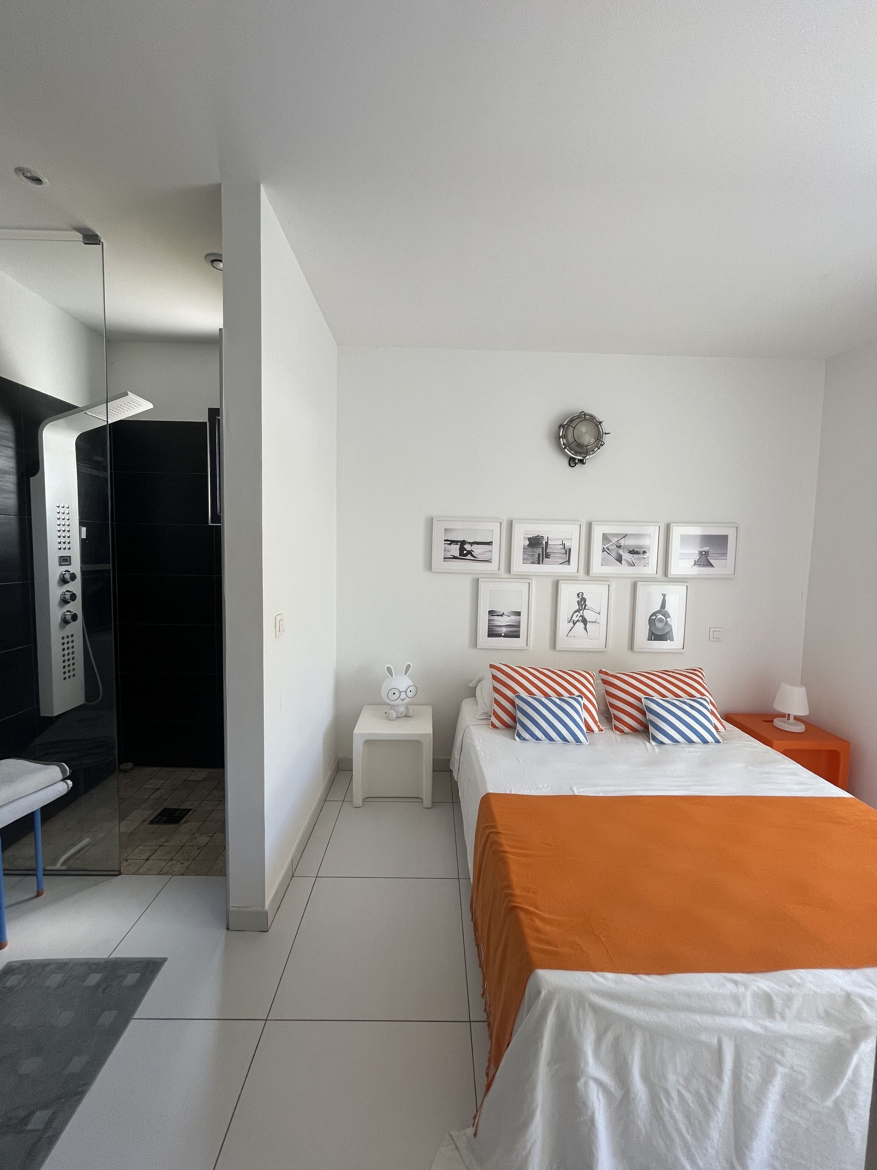 A modern hotel room with a bed, a nightstand, a small table with a decorative item, a shower area, and a wall with framed pictures.