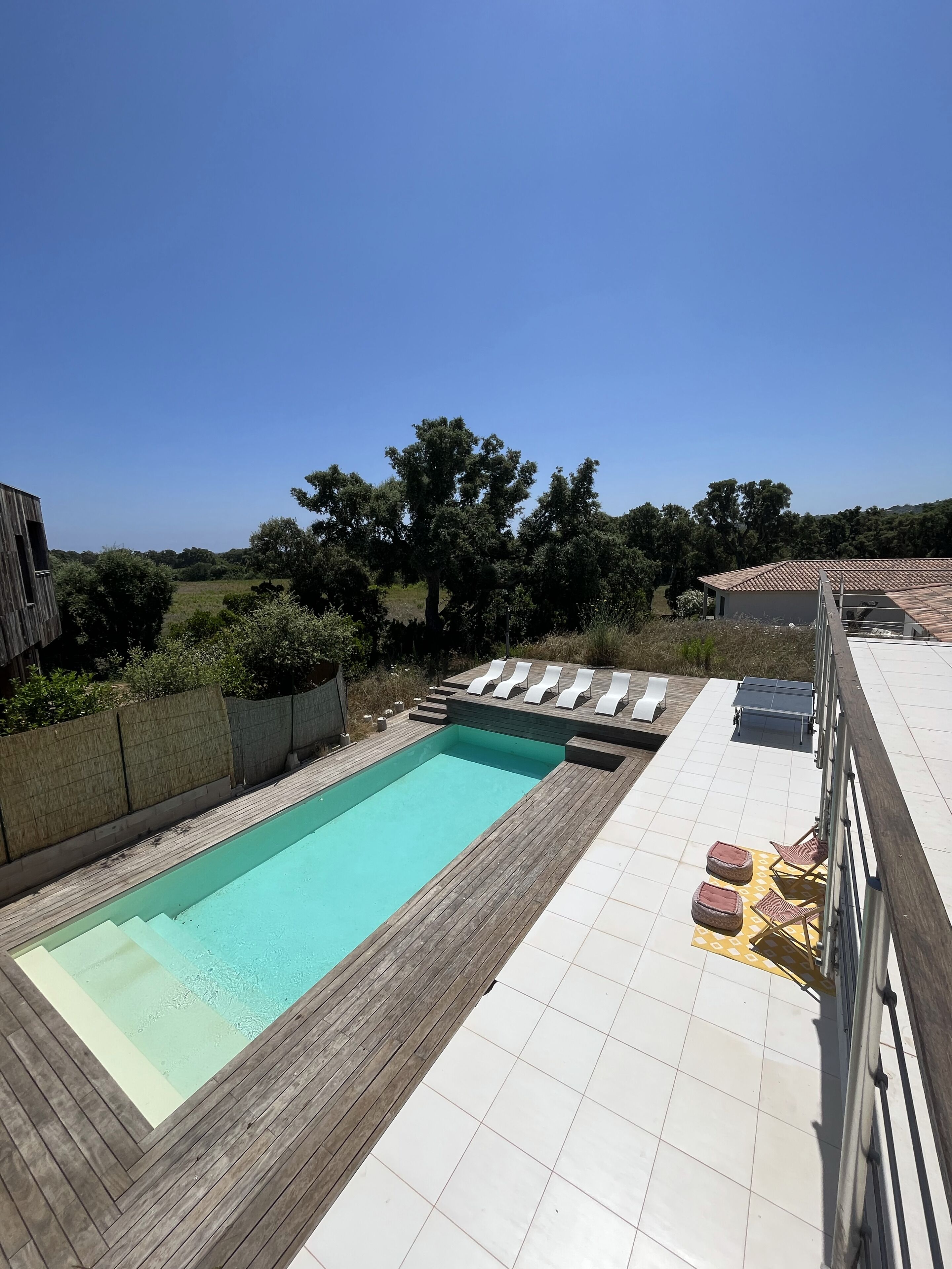 A rectangular swimming pool with a wooden deck surrounding it, a sun terrace with lounge chairs, and a view of the surrounding landscape.
