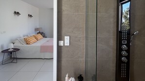 Un baño moderno con cabina de ducha acristalada, una mesita de noche con artículos de aseo y una cama en la habitación contigua.