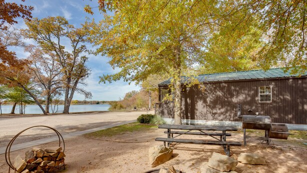 A rustic cabin with a picnic table and fire pit, surrounded by trees and a lake view. - Reservoir & Dock Access: Pet-Friendly Kerens Cabin (Kerens)