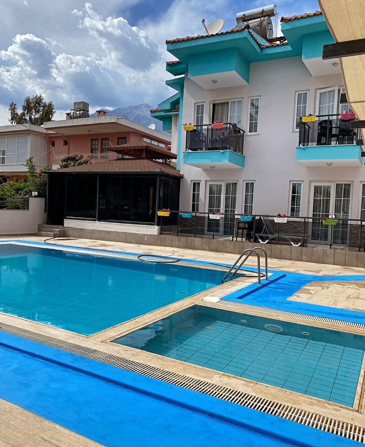 Una piscina con un borde de azulejos azules, una zona de estar cubierta y un edificio con balcones y ventanas al fondo.