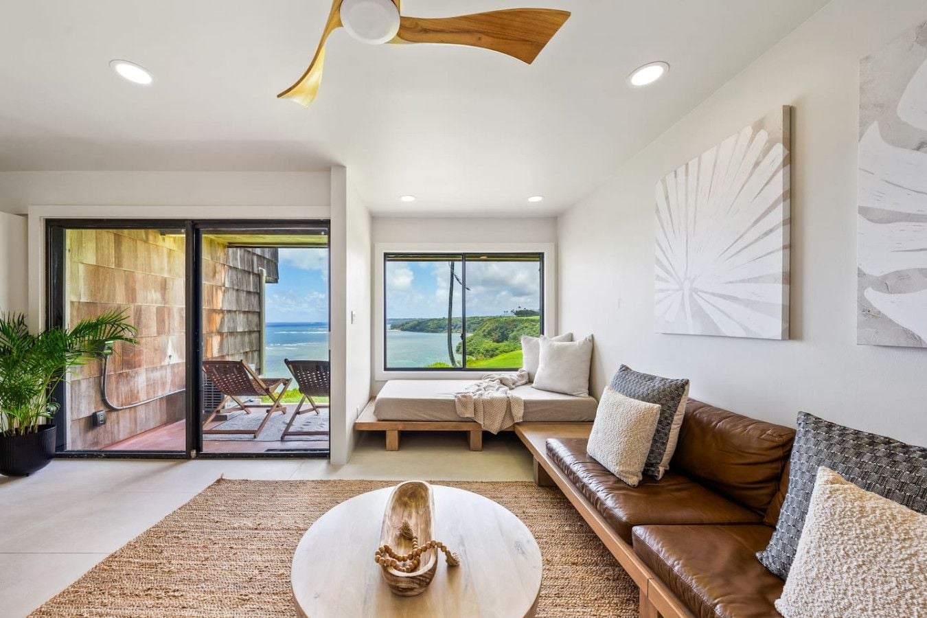 A modern living room with a large window offering a view of the sea, a cozy seating area, and a ceiling fan.