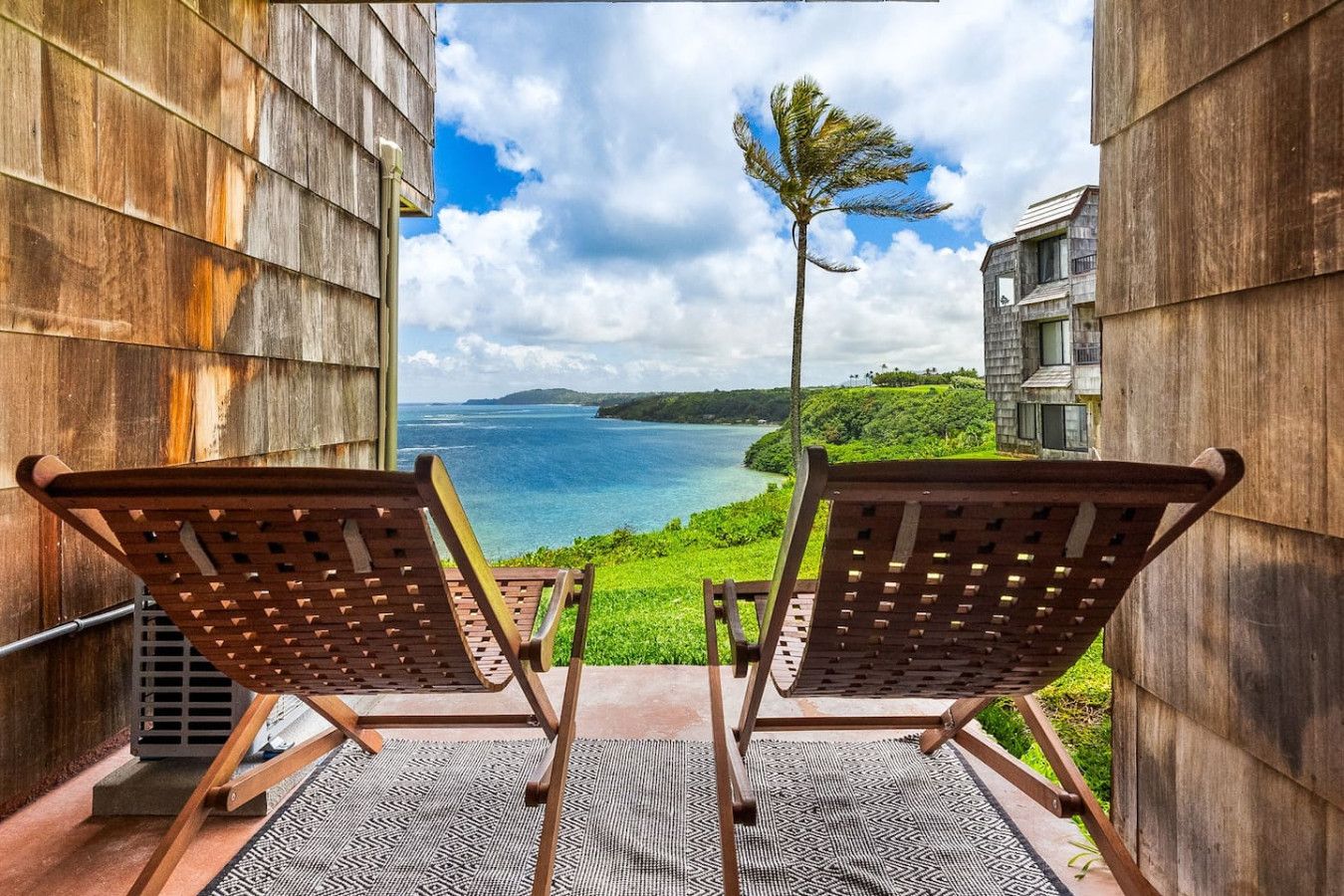 Two wooden lounge chairs facing a scenic ocean view from a balcony with a patterned rug.