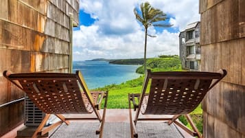 Two wooden lounge chairs facing a scenic ocean view from a balcony with a patterned rug.