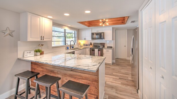 A kitchen with white cabinets, a granite countertop, wooden stools, a built-in oven, and a microwave. - Nautical Nook Pool Home (Brandon)