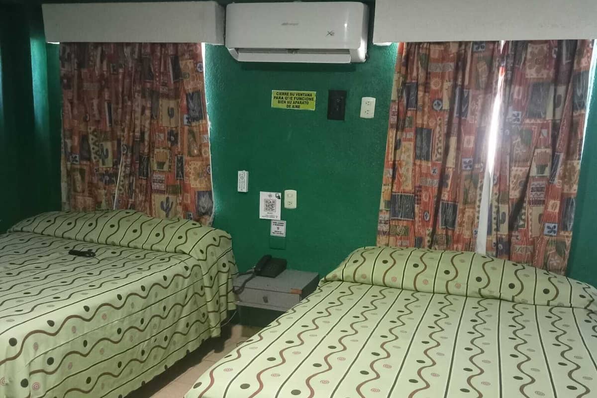 A room with two beds, each with patterned bedding, a small table, and an air conditioning unit.