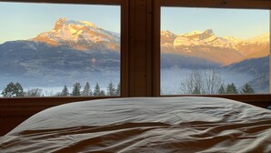 Una habitación con una cama y dos ventanas que ofrecen vistas a montañas nevadas.