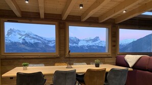 Un comedor con mesa de madera, cuatro sillas y un sofá. Hay tres ventanas amplias con vistas a las montañas.