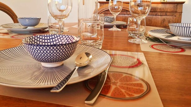 Una mesa de comedor dispuesta con platos, tazones y vasos, con una silla de madera visible al fondo.