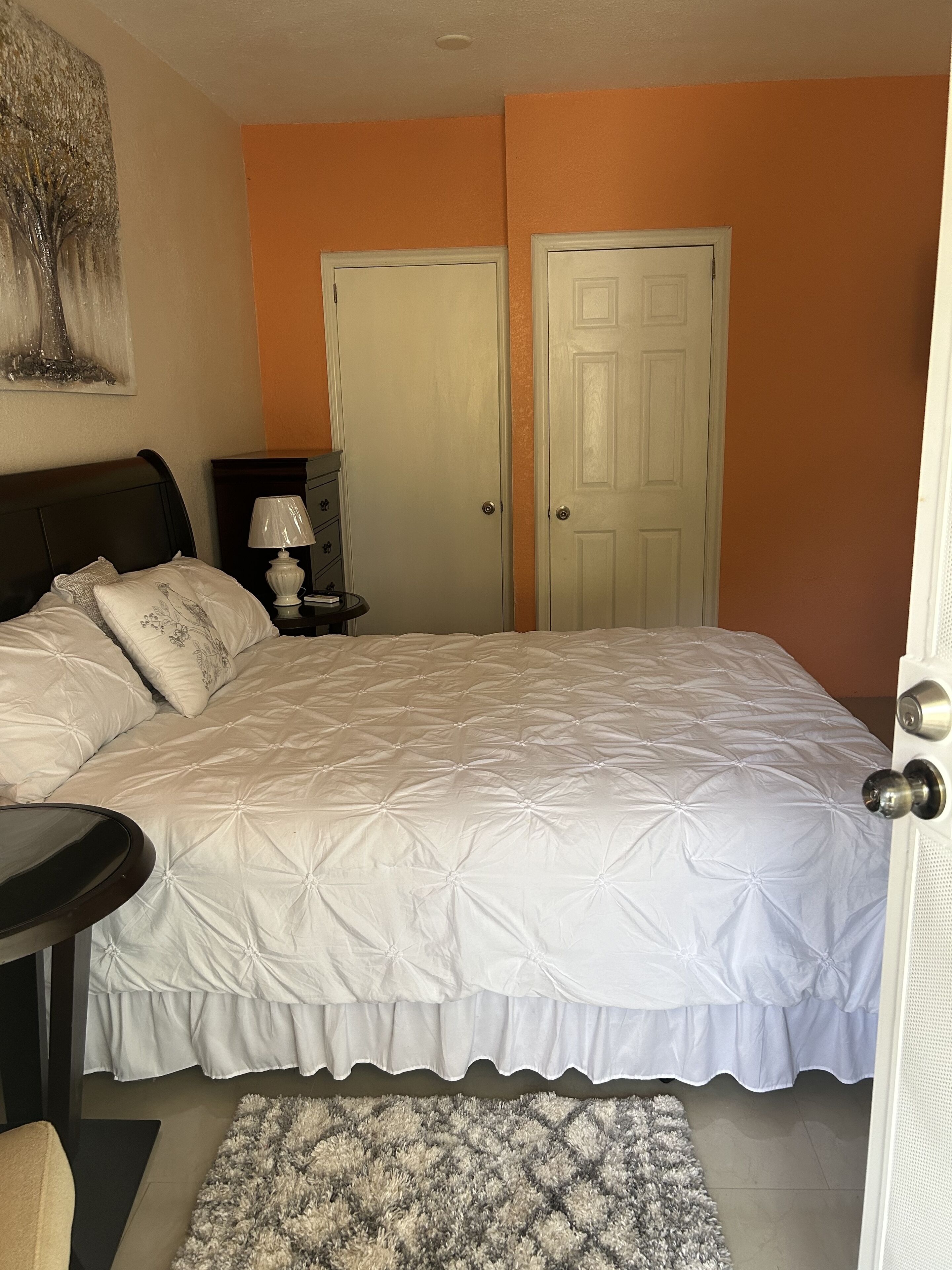 A bedroom with a large bed, two doors, a nightstand, a lamp, and a small rug.