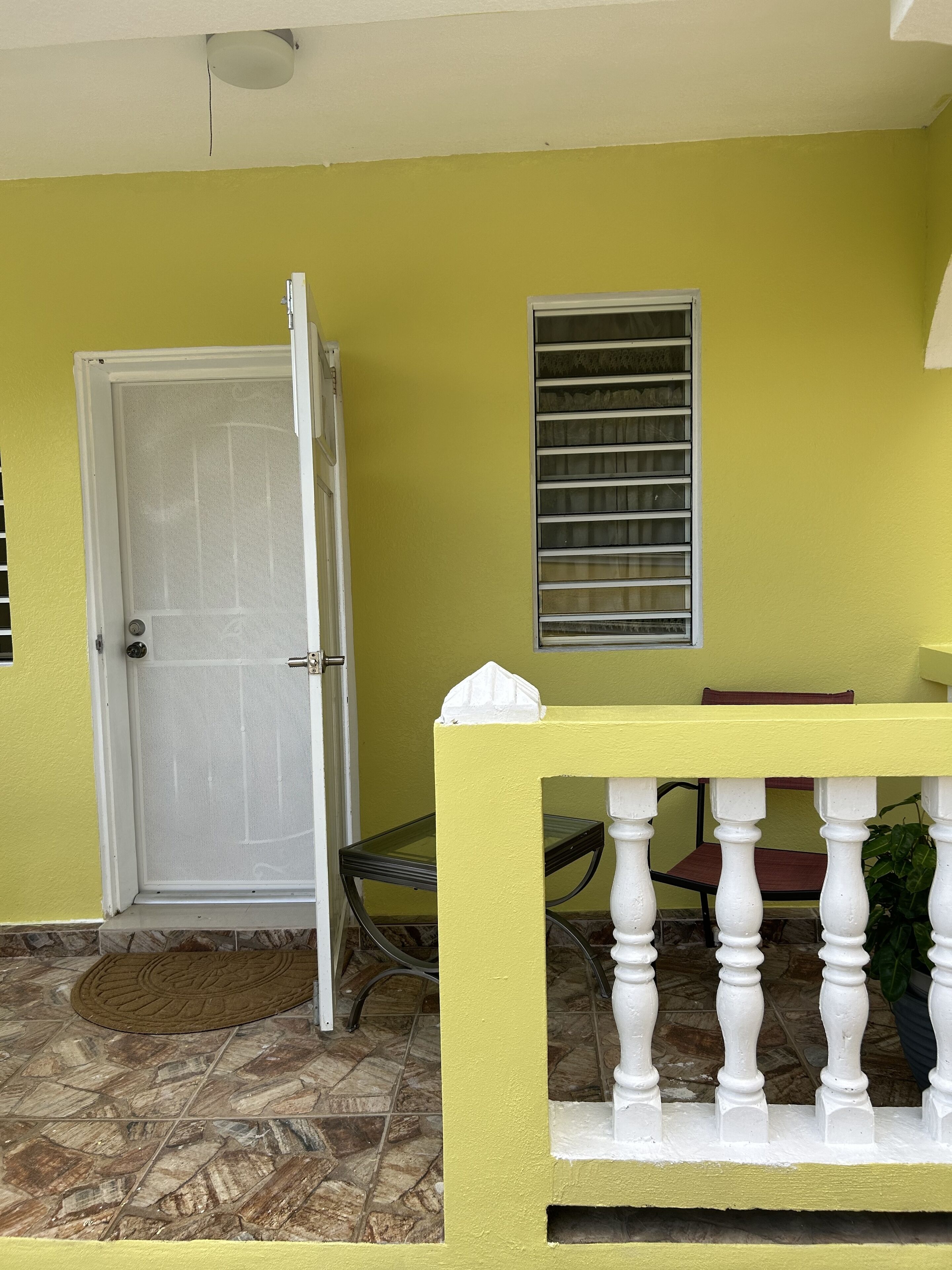 Un mur extérieur jaune avec une porte blanche, une fenêtre dotée de stores horizontaux et une petite table avec une balustrade blanche.