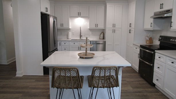 Une cuisine moderne avec des meubles blancs, un îlot central doté de tabourets, des appareils en acier inoxydable et une chaise en bois.