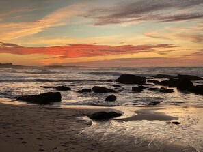 Ein Strand mit Felsen im Vordergrund und einem Sonnenuntergang im Hintergrund.