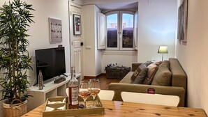 A cozy living room with a TV, a dining table with a bottle and glasses, a sofa, and a large window with a view.