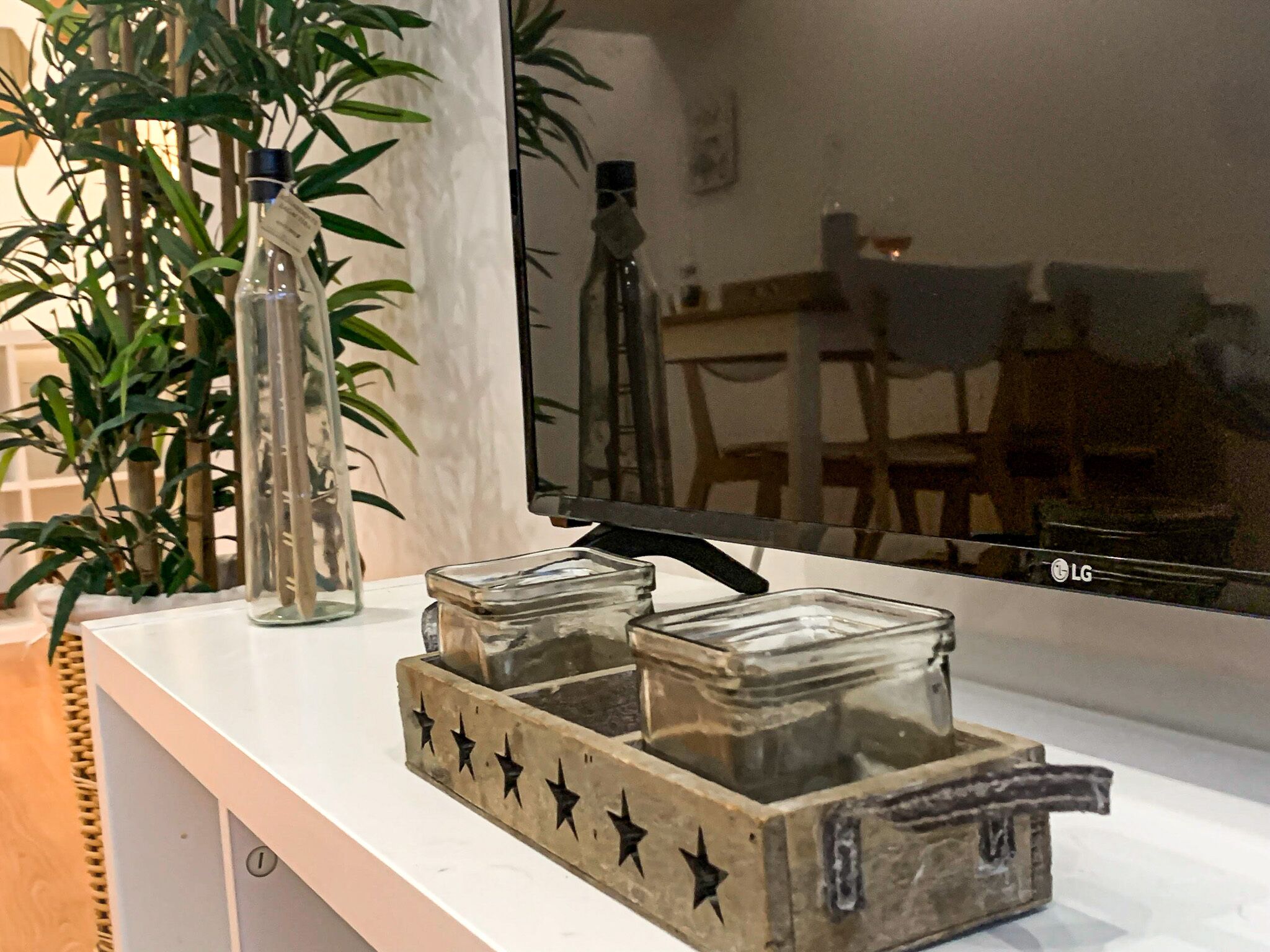 A kitchen counter with a glass container holding a bottle, a decorative wooden tray with star patterns, and a reflection of a dining area in a mirror.