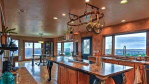 A spacious kitchen with a large island, wooden cabinetry, and a view of the outdoors through large windows. - Canyonside Retreat - Park-like setting, Panoramic Views with Hot tub & Firepit!! (Buhl)