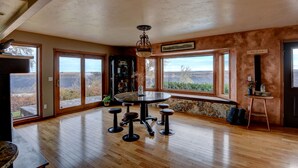 A spacious room with a wooden floor, large windows offering a view of the outdoors, a round table with stools, and a ceiling fan. - Canyonside Retreat - Park-like setting, Panoramic Views with Hot tub & Firepit!! (Buhl)