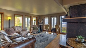 A spacious living room with a leather sofa, armchairs, a coffee table, and a fireplace. Large windows provide a view of the outdoors. - Canyonside Retreat - Park-like setting, Panoramic Views with Hot tub & Firepit!! (Buhl)