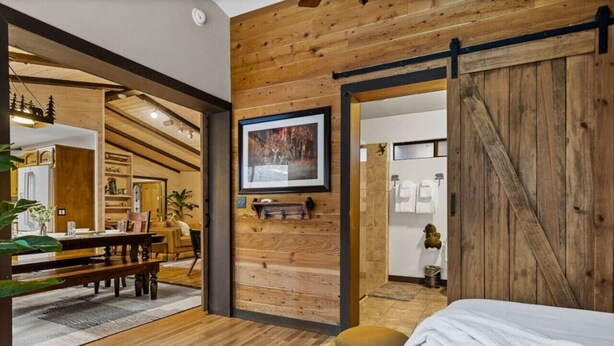 A modern room with wooden walls, a bed, a dining area, and a kitchen visible through an open door. - Ritz-Craterton|Spa + BBQ + Game Room + EV + Pups (Big Bear Lake)