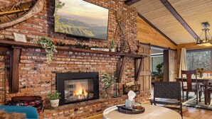 A cozy living room with a brick fireplace, a large painting, and a dining area with wooden furniture. - Ritz-Craterton|Spa + BBQ + Game Room + EV + Pups (Big Bear Lake)