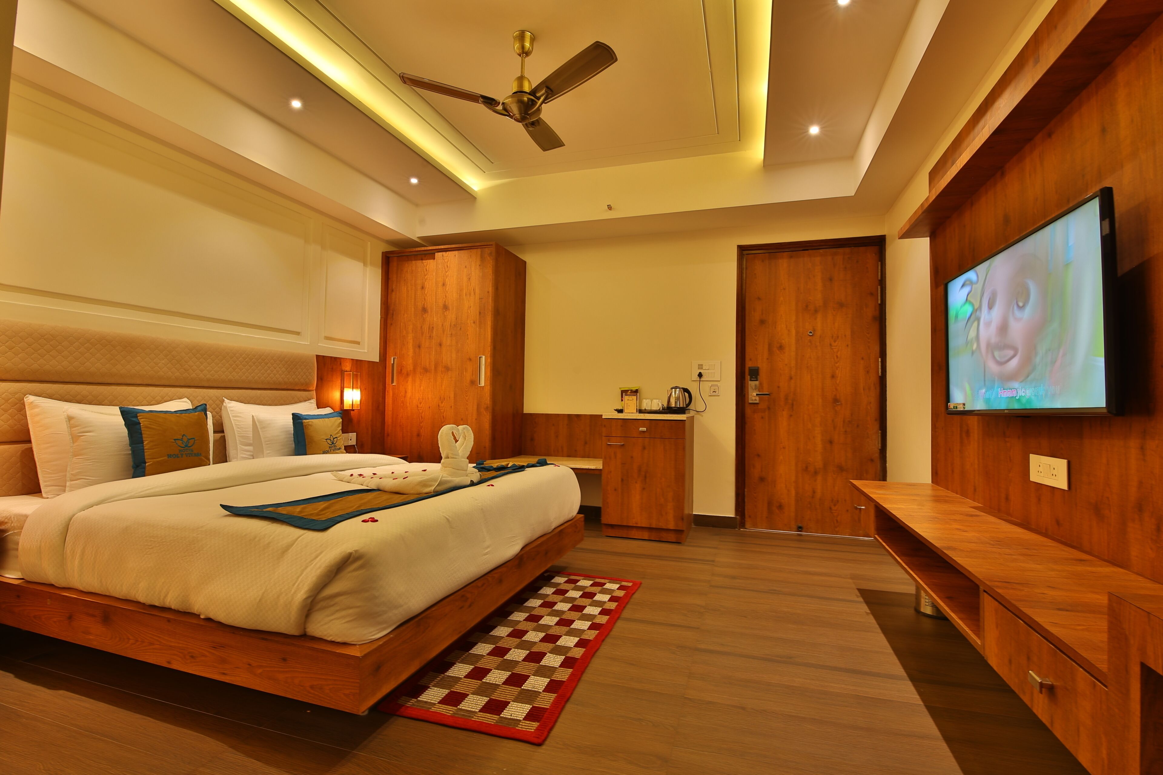 A modern bedroom with a large bed, wooden wardrobe, a TV, and a ceiling fan.