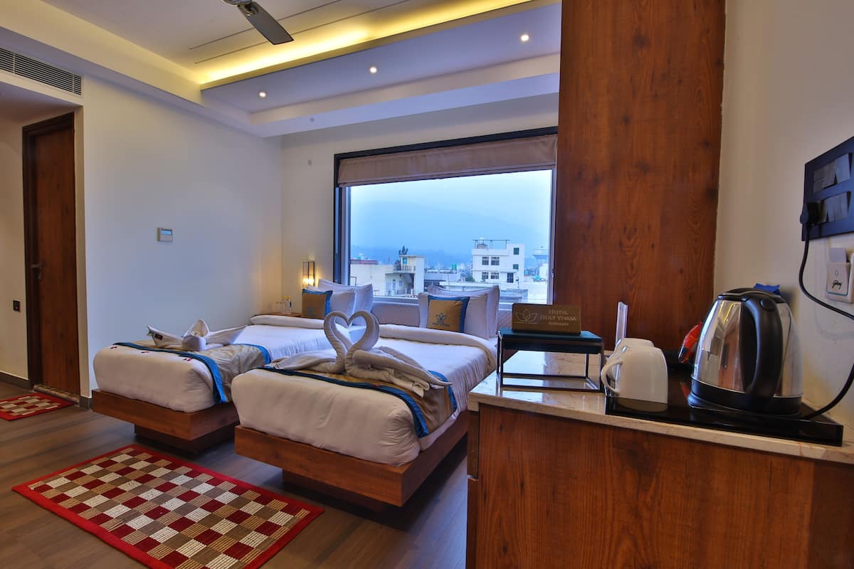 A hotel room with two beds, a large window, a wooden desk with a kettle, and a red and beige rug.