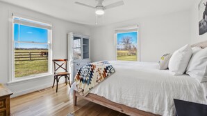 A bedroom with a bed, a chair, a desk, and a ceiling fan. There are two windows with views of a grassy field and a fence.
