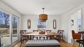 A dining room with a large wooden table, six chairs, a bench, and a hanging basket. There are two windows with a view of a deck and trees.