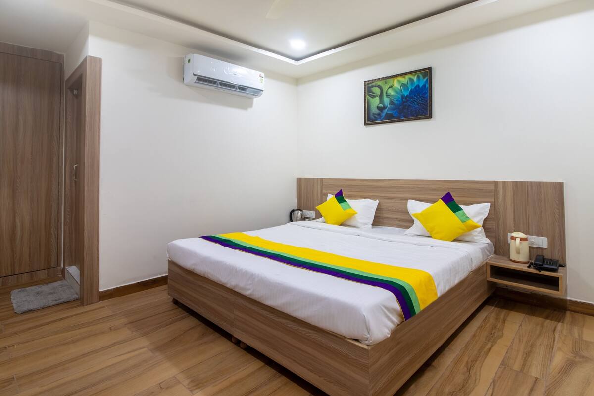 A neatly arranged bedroom with a wooden bed, white bedding, a colorful blanket, a bedside table with a phone and a cup, and an air conditioning unit on the wall.