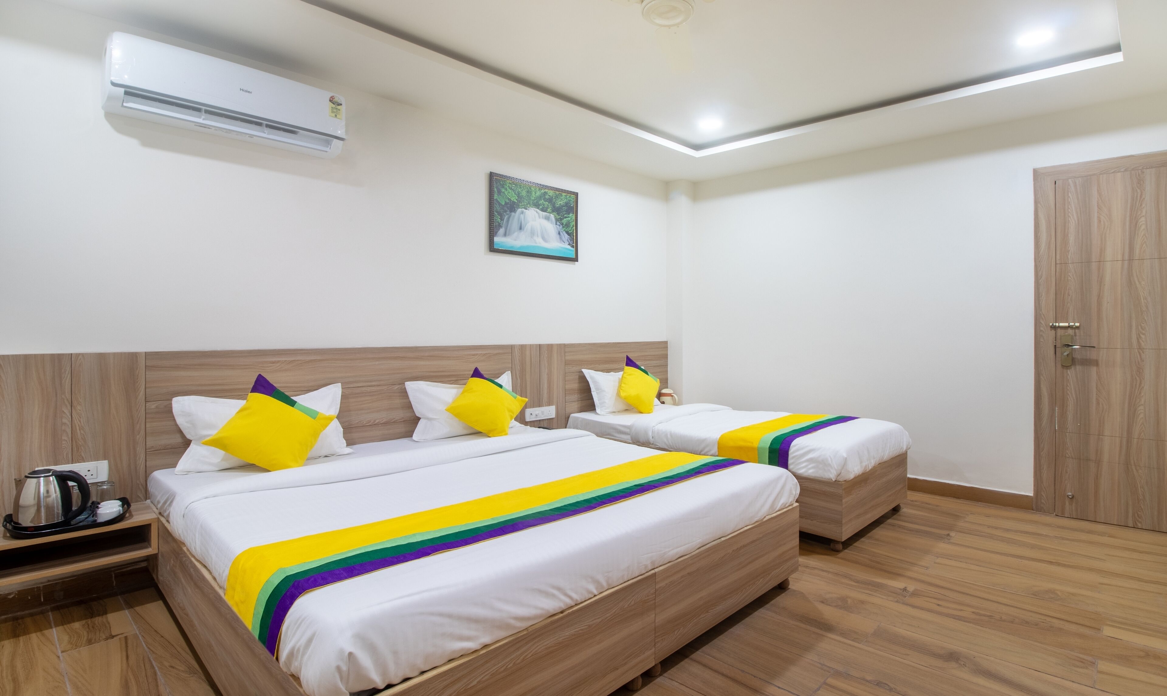 A hotel room with two beds, a wooden floor, a painting on the wall, and an air conditioning unit.