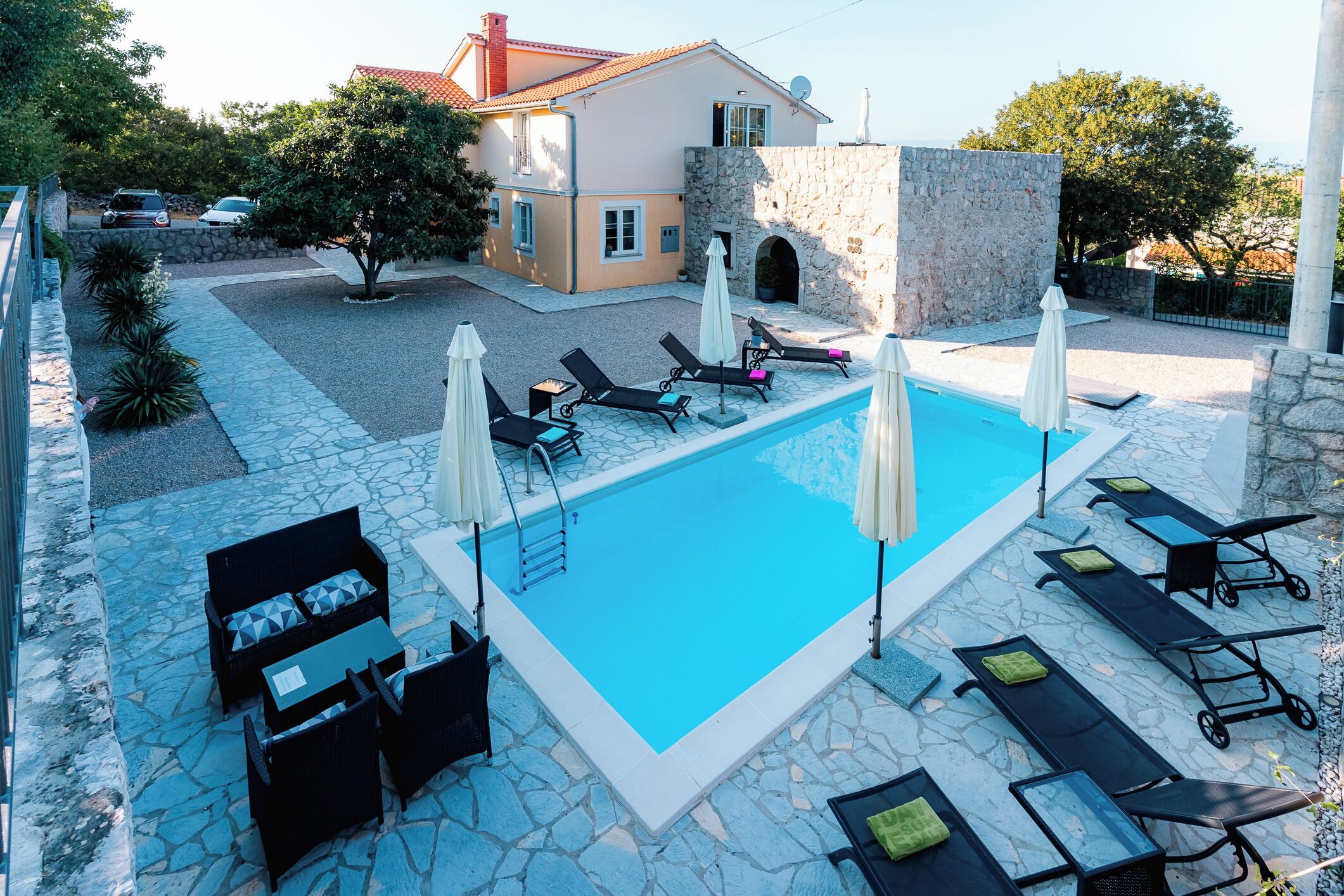 A swimming pool with lounge chairs and umbrellas, surrounded by stone tiles and a stone wall.