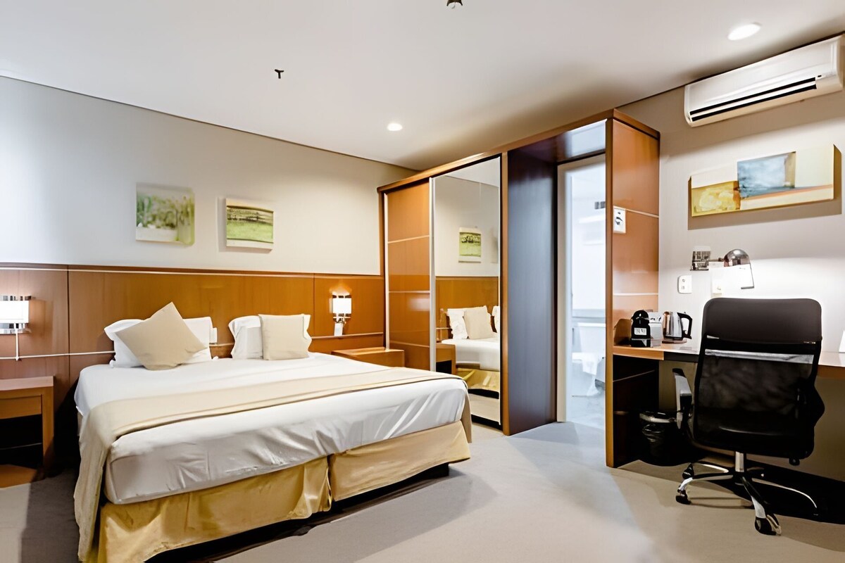 Habitación de hotel con una cama grande, un escritorio, una silla, equipo de aire acondicionado y obras de arte en las paredes.