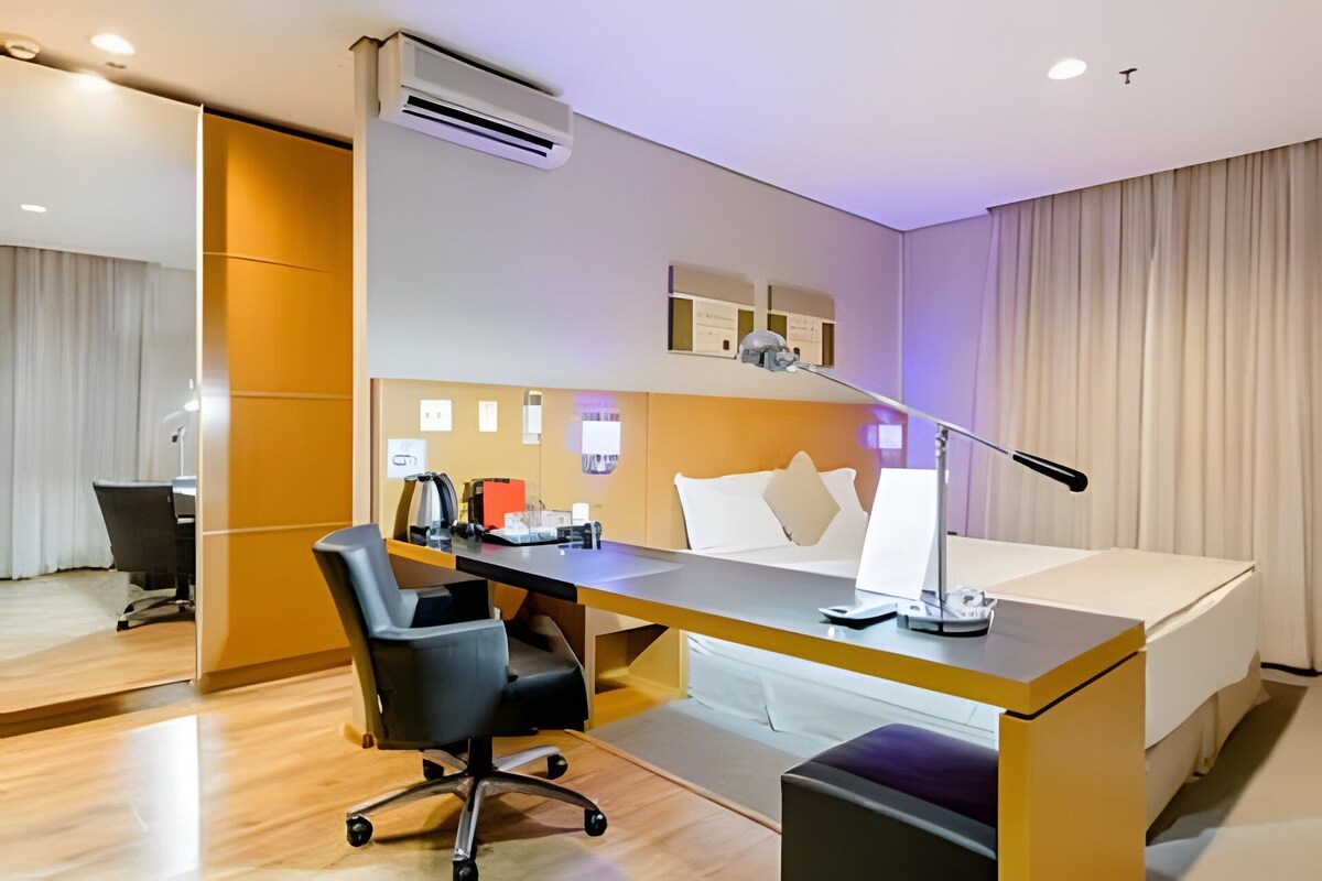 Una habitación de hotel con cama, escritorio, silla y unidad de aire acondicionado.