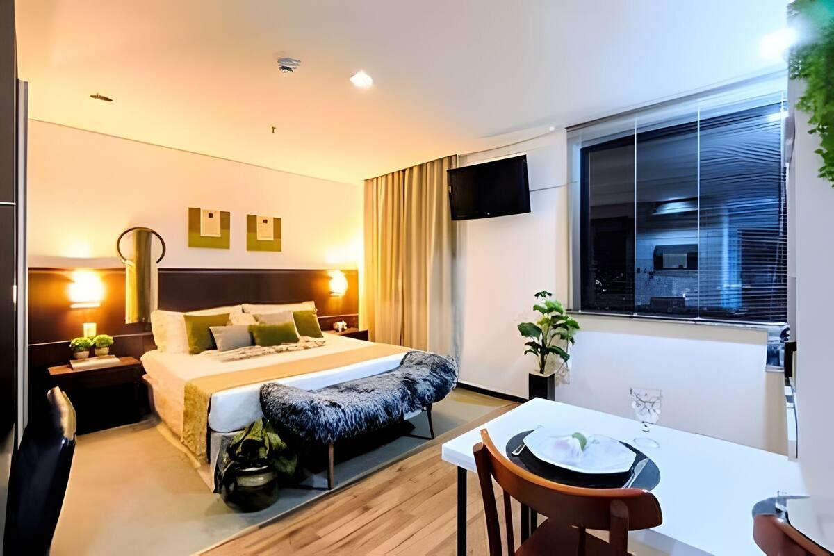 Ein modernes Hotelzimmer mit Bett, Nachttisch, Lampe, Fernseher, Fenster und einer Essecke mit Tisch und Stühlen.
