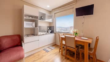 Cozinha compacta com armários brancos, micro-ondas e área de refeições com mesa e cadeiras. Uma cadeira vermelha é visível, e uma TV está montada na parede.
