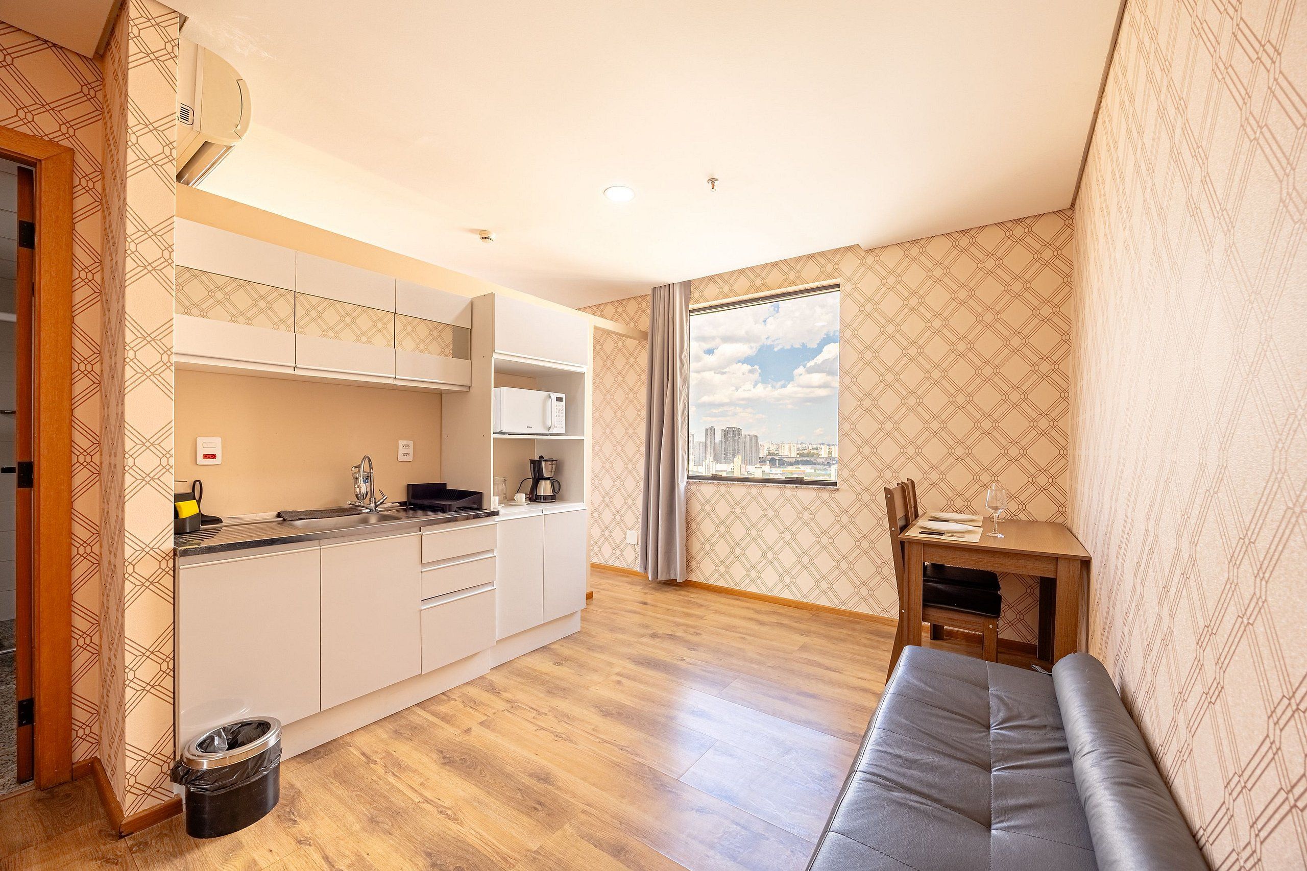 A compact living space with a kitchenette, a dining area, and a large window offering a city view.
