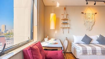 A bedroom with a bed, a red chair, a table with a lamp, a macrame wall hanging, and a city view.