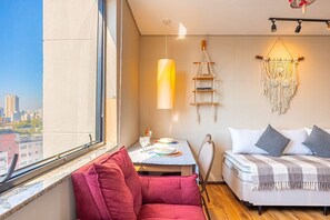A bedroom with a bed, a red chair, a table with a lamp, a macrame wall hanging, and a city view. - Flat Osasco Trade (Osasco)
